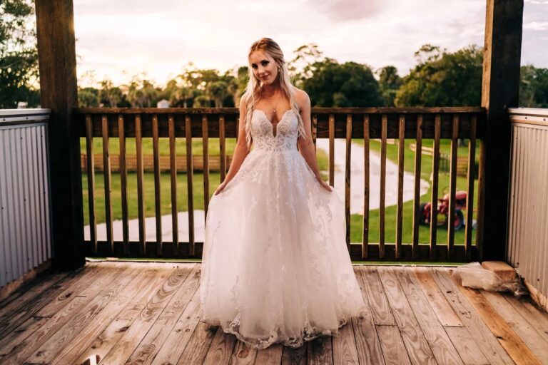 A beautiful bride in a lace wedding gown poses on a wooden deck overlooking a scenic South Florida ranch wedding venue, with lush greenery and a winding pathway in the background.