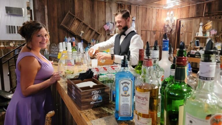 A smiling bridesmaid in a purple dress stands at a rustic bar while a bartender in a vest prepares drinks, capturing a lively moment at a ranch wedding in South Florida.