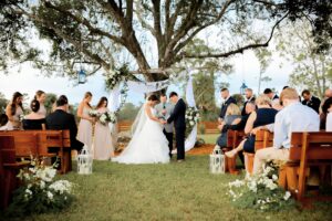 Outdoor wedding ceremony setup near Palm Beach at Rockin H Ranch, featuring rustic wooden benches, lush greenery, and a scenic countryside backdrop.