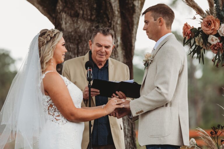 Bride and groom exchange vows during an outdoor wedding ceremony at Rockin H Ranch near Palm Beach, officiated beneath a tree with floral decor.