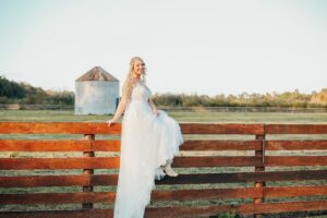 A smiling bride in a flowing white gown sits elegantly on a wooden fence at a South Florida ranch wedding venue, with a picturesque barn and open fields in the background.