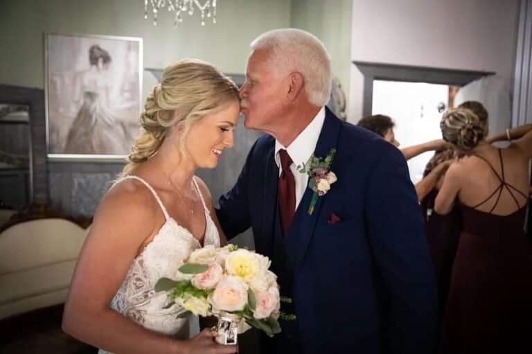 The bride holding a floral bouquet smiles as her father kisses her forehead in a touching moment during a South Florida ranch wedding.