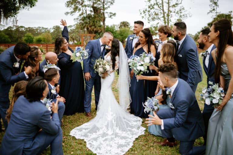 The bride and groom kissing, surrounded by their bridal party dressed in blue and holding floral bouquets, set in a picturesque South Florida ranch wedding.