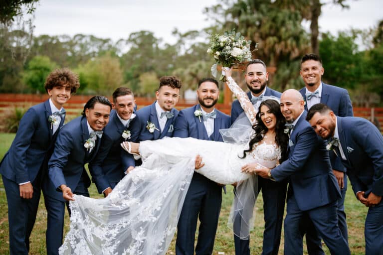 Group photo of a bride in a lace wedding dress being lifted by groomsmen in blue suits, captured at an outdoor wedding venue near Palm Beach, Florida.