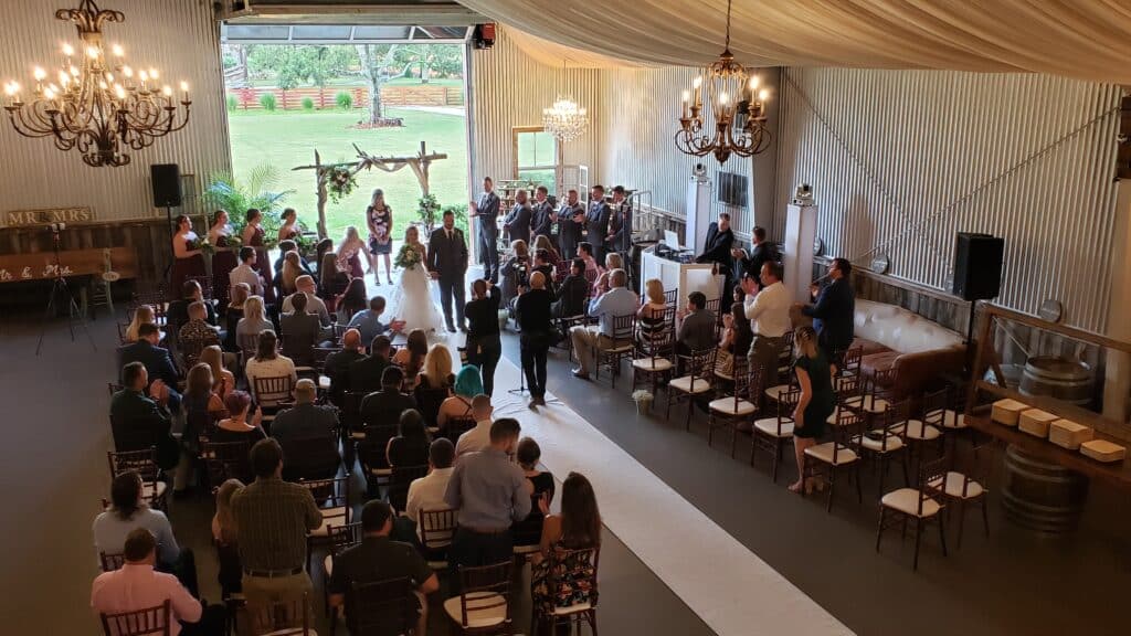 Indoor wedding ceremony with guests seated, chandeliers overhead, and a view of the outdoor greenery through open doors at a venue near Palm Beach, Florida.