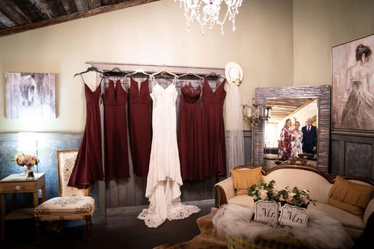 A white bridal gown and burgundy bridesmaid dresses hanging in a rustic ranch-style bridal suite, decorated with vintage furniture, soft lighting, and elegant wedding accents in South Florida.