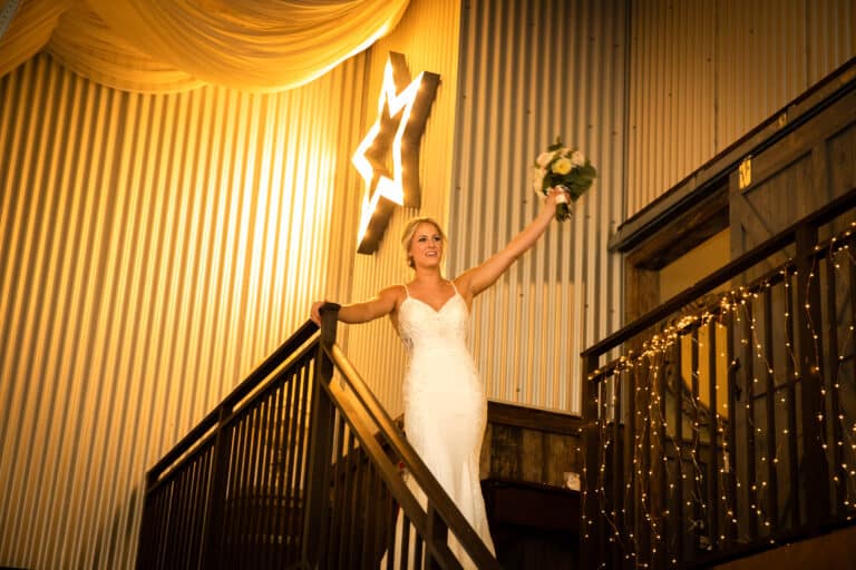 A bride in an elegant white gown stands at the top of a wooden staircase, holding her bouquet high in celebration at a rustic ranch wedding venue in South Florida, illuminated by warm fairy lights and a glowing star sign.