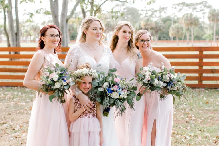 The bride, bridesmaids, and flower girl smiling and holding floral bouquets in a serene South Florida ranch wedding setting with a wooden fence backdrop.